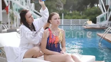 母亲和女儿坐在游泳池旁的休息室里。 母亲梳女儿`头发。